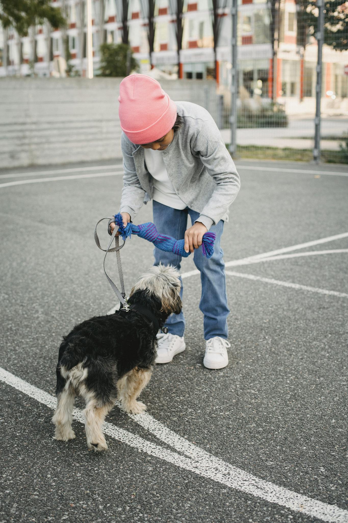 A child in a pink hat trains a dog on a city sports ground during the day.